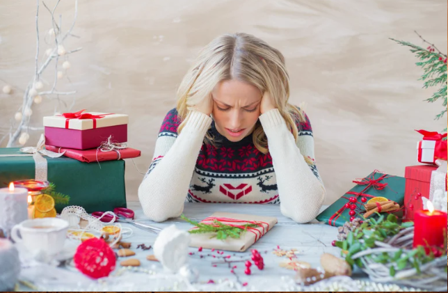 A woman sits at a table surrounded by Christmas presents, looking overwhelmed by holiday stress.