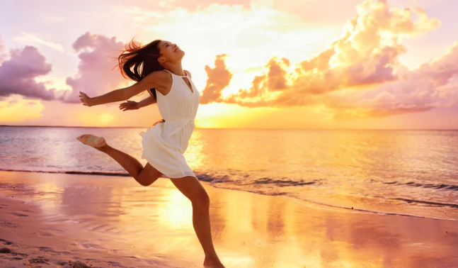 A woman in a white dress joyfully jumps on the beach as the sun sets, casting a warm glow over the scene.